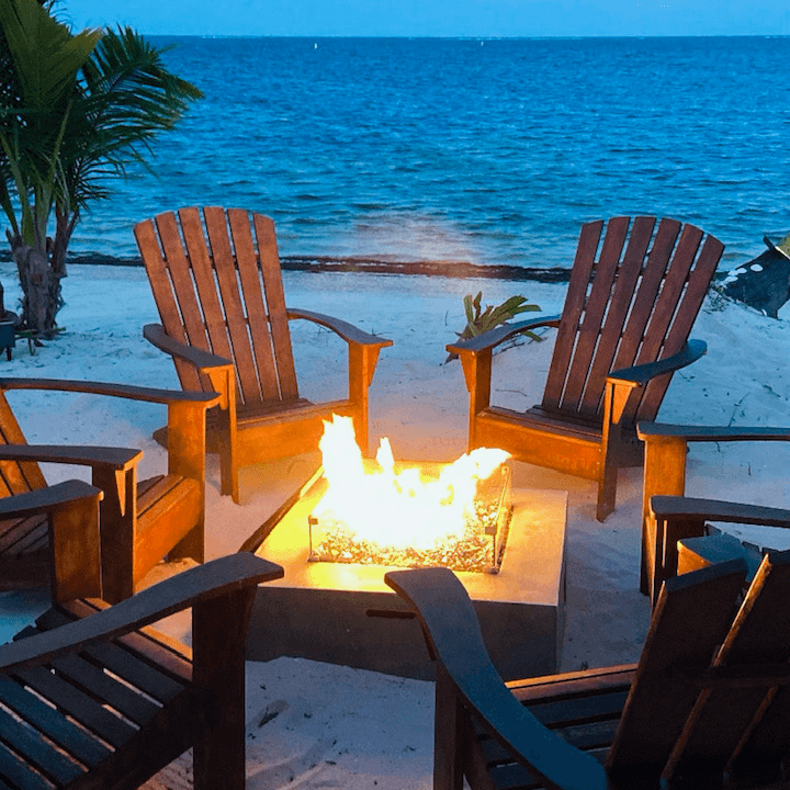 A circular arrangement of wooden chairs surrounding a glowing fire pit on a sandy beach by the water.