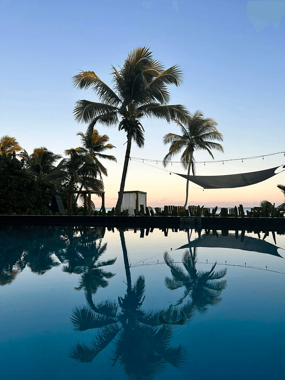 Silhouetted palm trees and a hammock reflect in a tranquil pool at sunset.