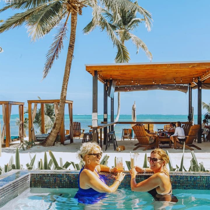 Two women enjoy drinks by a pool with a beachside view and palm trees in the background.